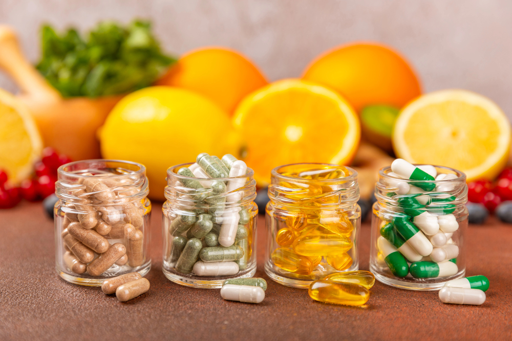 Four small glass jars filled with assorted capsules and softgel pills are displayed in front of sliced citrus fruits, berries, and leafy greens on a brown surface.