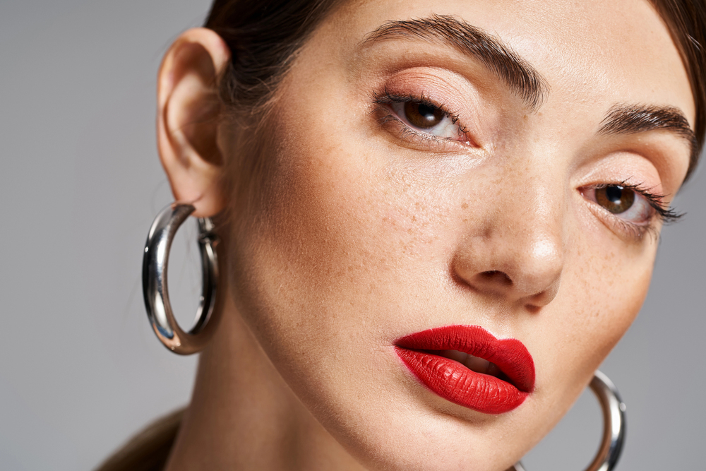 Close-up of a woman with fair skin, bold red lipstick, natural eye make-up, large silver hoop earrings, and brown hair pulled back, gazing confidently at the camera against a neutral background.