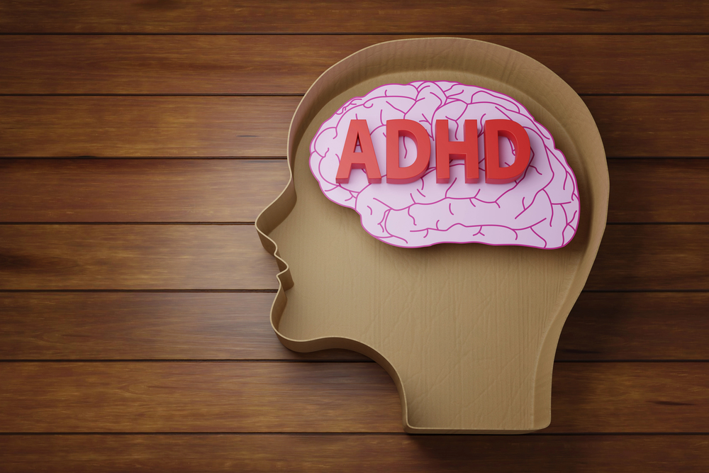 A wooden profile of a human head is shown with a pink brain illustration inside. The bold red letters ADHD are placed on top of the brain. The background is wooden planks.