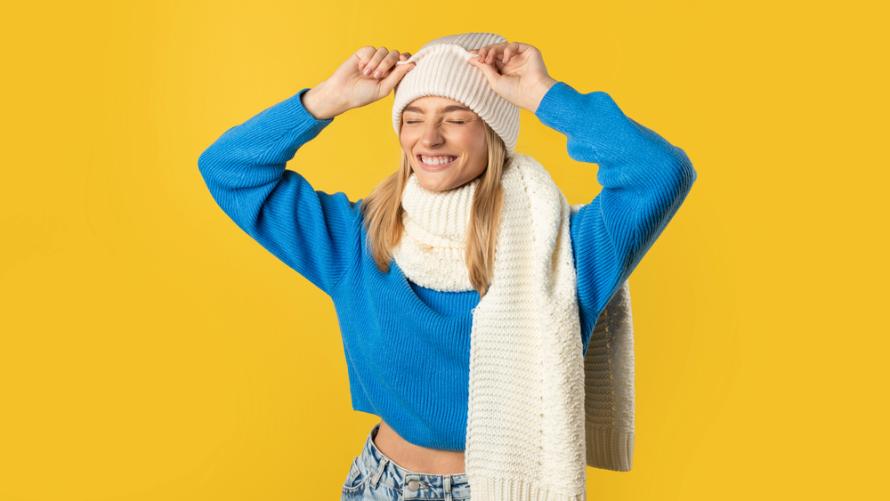 A smiling woman in a blue jumper, white knitted scarf, and white beanie adjusts her hat whilst standing against a bright yellow background. Her eyes are closed and she looks happy.