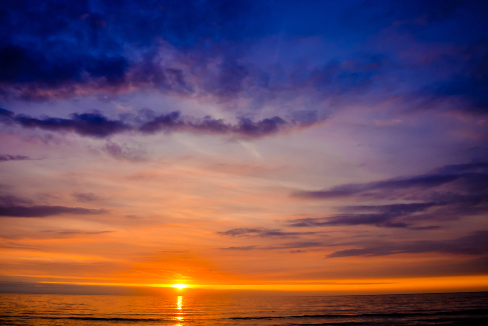 A vibrant sunset over the ocean, with the sun low on the horizon and the sky filled with colourful clouds in shades of purple, orange, and blue reflecting on the water.