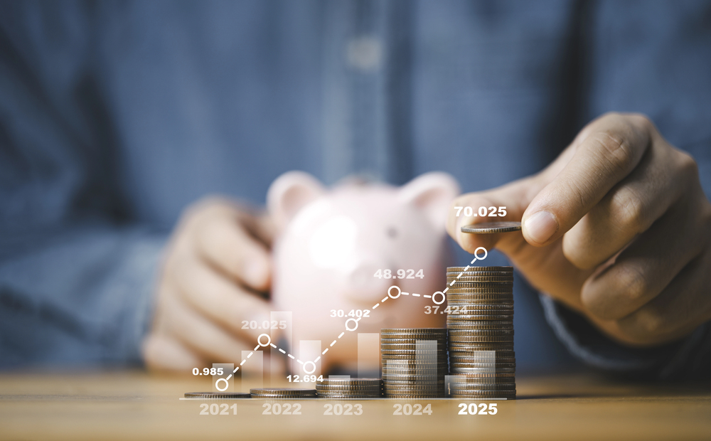 A person stacks coins in increasing order, with a pink piggy bank in the background. An upward graph with data points from 2021 to 2025 overlays the image, representing financial growth and savings.