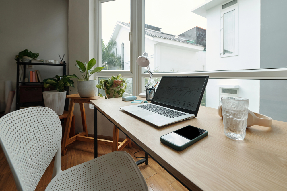 Minimalist home office with a laptop, mobile phone, glass of water, and potted plants on a wooden desk next to a large window overlooking neighbouring houses. White chairs are positioned at the desk.
