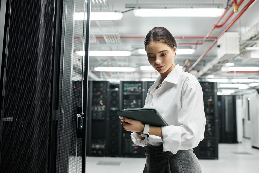 A woman in business attire stands in a server room, holding a tablet and looking at it thoughtfully. Tall racks of servers and cables are visible in the background.