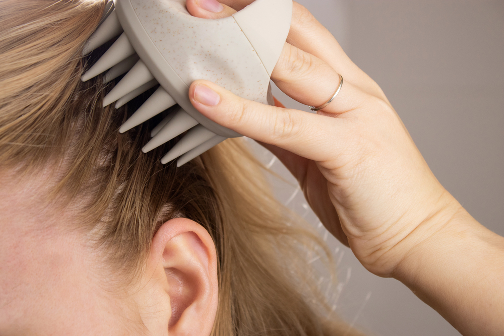 A person uses a scalp massager with soft silicone bristles on the side of their head, holding their blond hair back with one hand and wearing a thin ring on their finger.