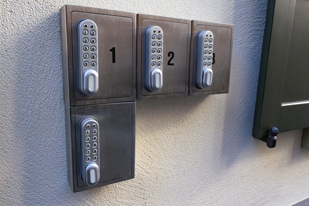 Four metal lockboxes with numbered keypads (1, 2, 3, 4) are mounted on a light-coloured wall near a green window shutter. Each lockbox has a combination dial for secure access.