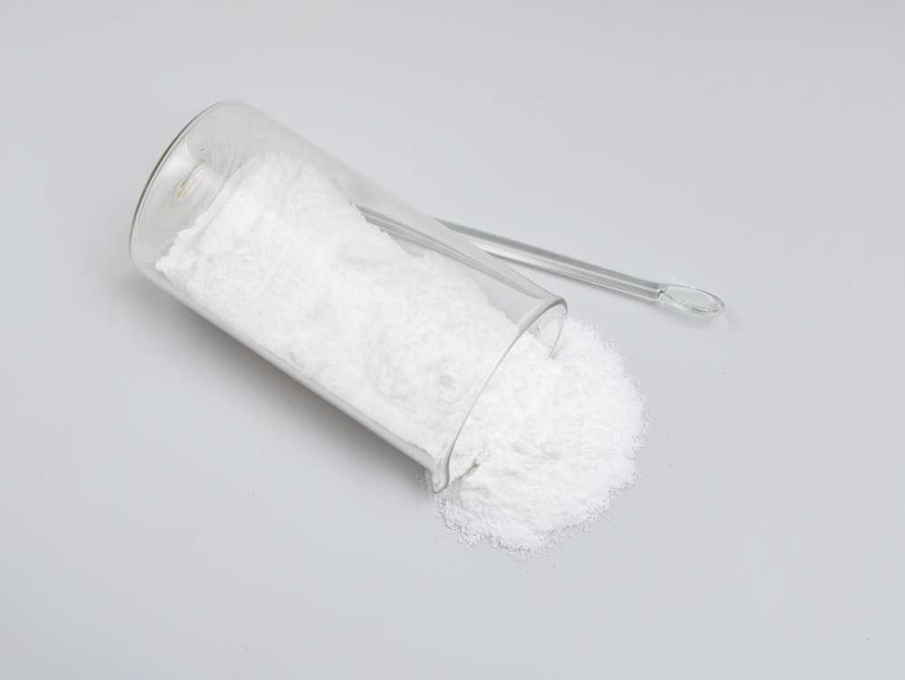 A glass beaker tipped over, spilling a pile of fine white powder onto a white surface, with a glass stirring rod lying next to it.