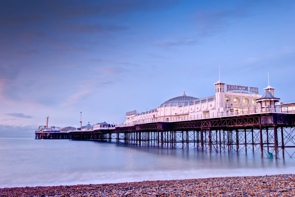 Brighton Pier extends over calm sea water at dusk, illuminated by soft lights, with a pebble beach in the foreground and a pastel sky above. The pier's sign and structure are clearly visible.