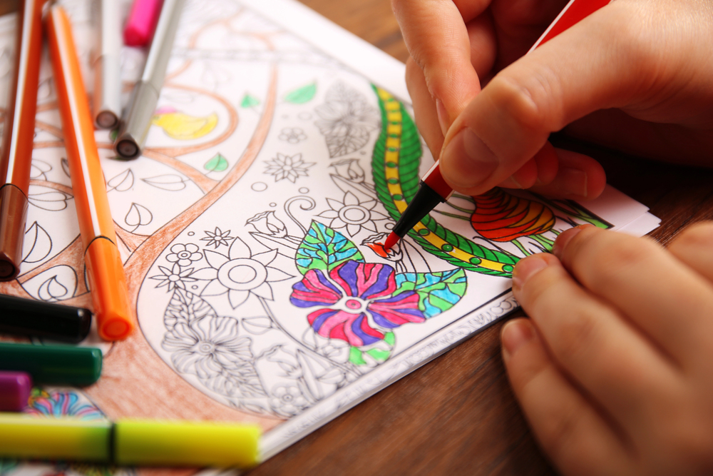 A person uses coloured pens to fill in a detailed floral colouring book page, with several coloured markers lying nearby on a wooden surface.