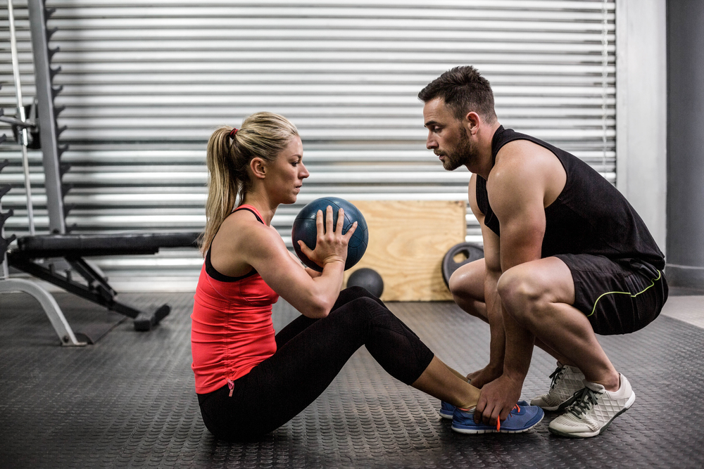 A woman in sportswear sits holding a medicine ball while doing sit-ups. A man in a black vest and shorts kneels in front of her, holding her feet to support her workout in a gym setting.