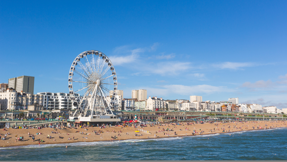 A large Ferris wheel stands near a busy beach with many people relaxing on the sand. City buildings and flats line the waterfront under a clear blue sky.