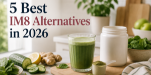 A glass of green smoothie, a white jar, and fresh ingredients like cucumber, lemon, ginger, spinach, and apples on a kitchen worktop. Text reads: 5 Best IM8 Alternatives in 2026.