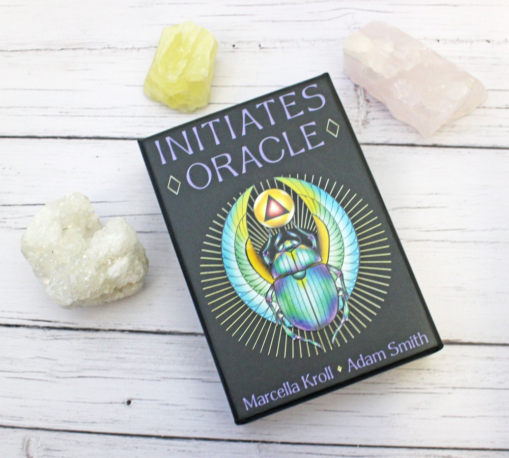 A box of Initiates Oracle cards by Marcella Kroll and Adam Smith lies on a white wooden surface, surrounded by three crystals: one yellow, one clear, and one pale pink. The box features a colourful scarab beetle design.