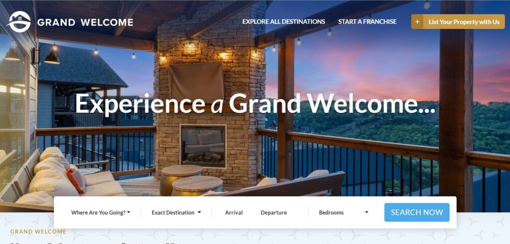 A cosy outdoor seating area with sofas and a fireplace on a covered deck overlooks mountains at sunset. Website text reads, “Experience a Grand Welcome…” with a search bar for booking holiday rentals.
