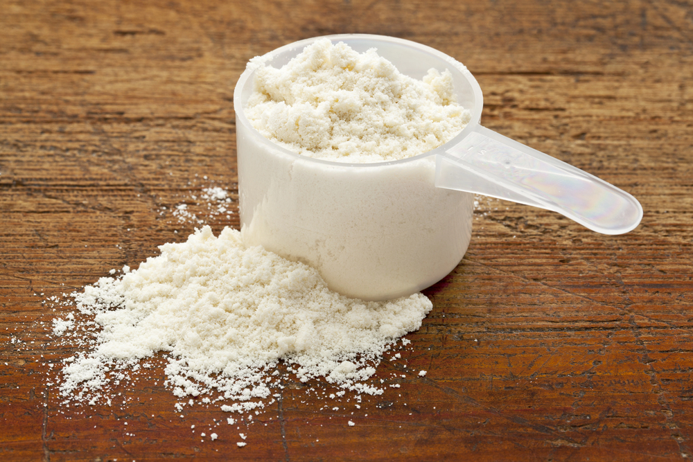 A plastic scoop filled with white protein powder sits on a wooden surface, with some powder spilt beside it.