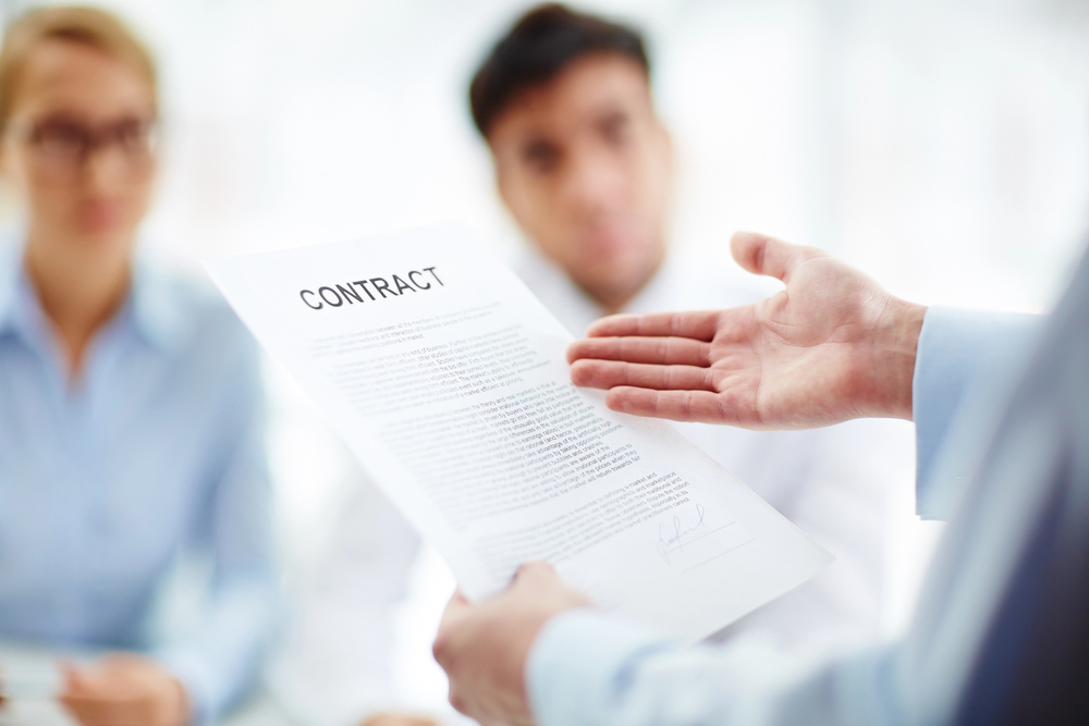A person holds out a contract and gestures whilst two people sit opposite across the table, watching and listening in a bright, professional setting.