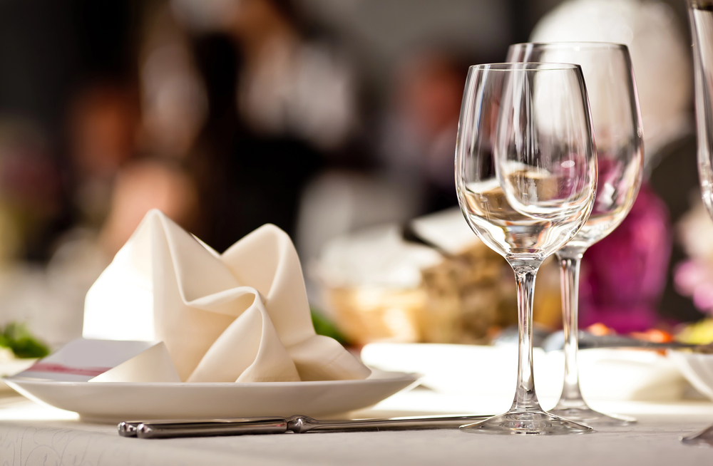 Two empty wine glasses stand beside a plate with a neatly folded white serviette on a table laid for a formal meal, with blurred background elements.