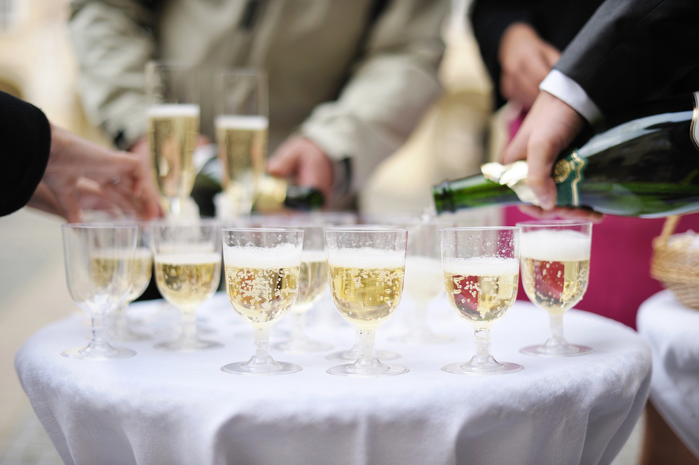 Several glasses of sparkling wine are arranged on a round table with a white tablecloth. People are pouring and picking up drinks, with hands and bottles visible in the scene. The background is blurred.
