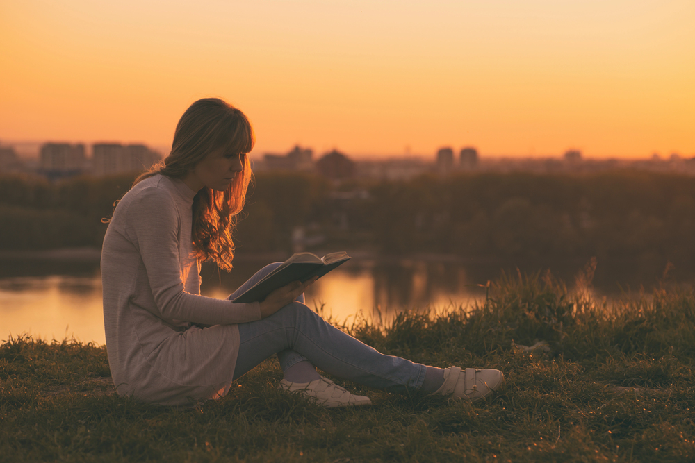 A young woman sits on grass near a body of water at sunset, reading a book. The sky is orange, and city buildings are visible in the distance.
