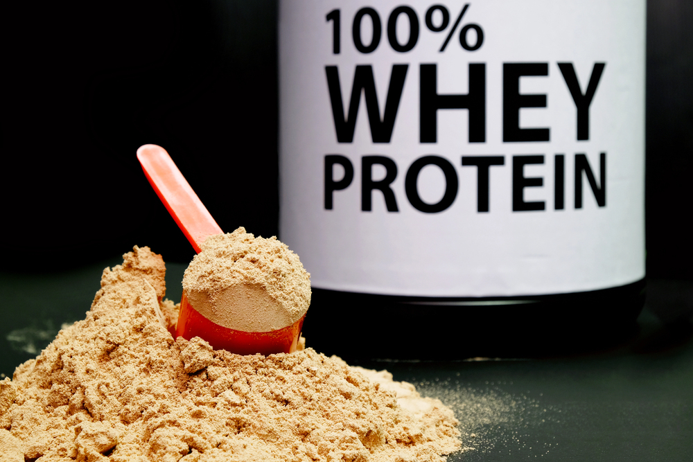 A red scoop filled with protein powder rests on a pile of powder in front of a large container labelled 100% Whey Protein on a black background.