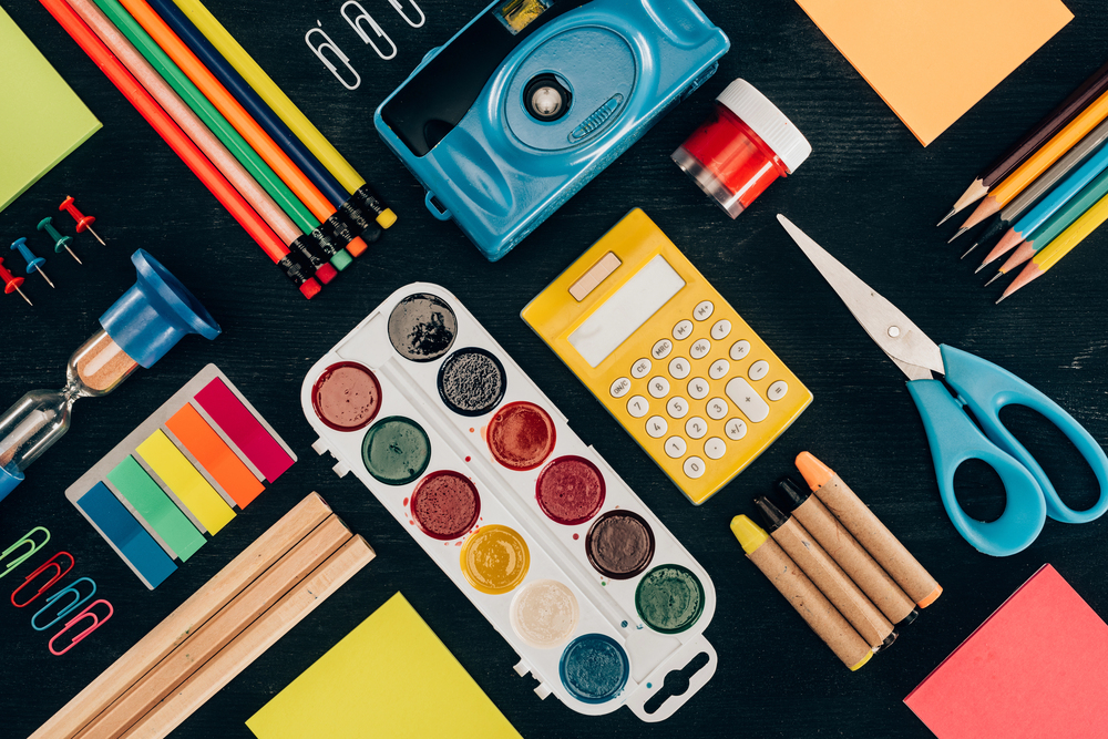 Assorted school supplies, including coloured pencils, markers, watercolour paints, scissors, a calculator, an hourglass, drawing pins, and paper, are neatly arranged on a dark surface.