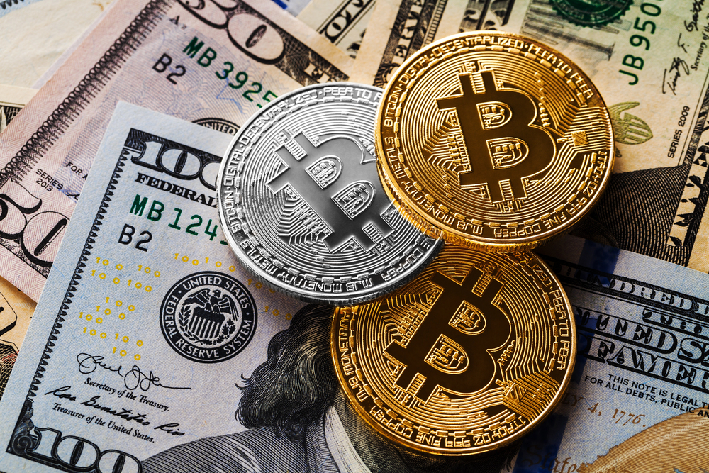 Three Bitcoin coins (two gold, one silver) are placed on top of various US dollar notes, including $5, $10, $20, $50, and $100 denominations.