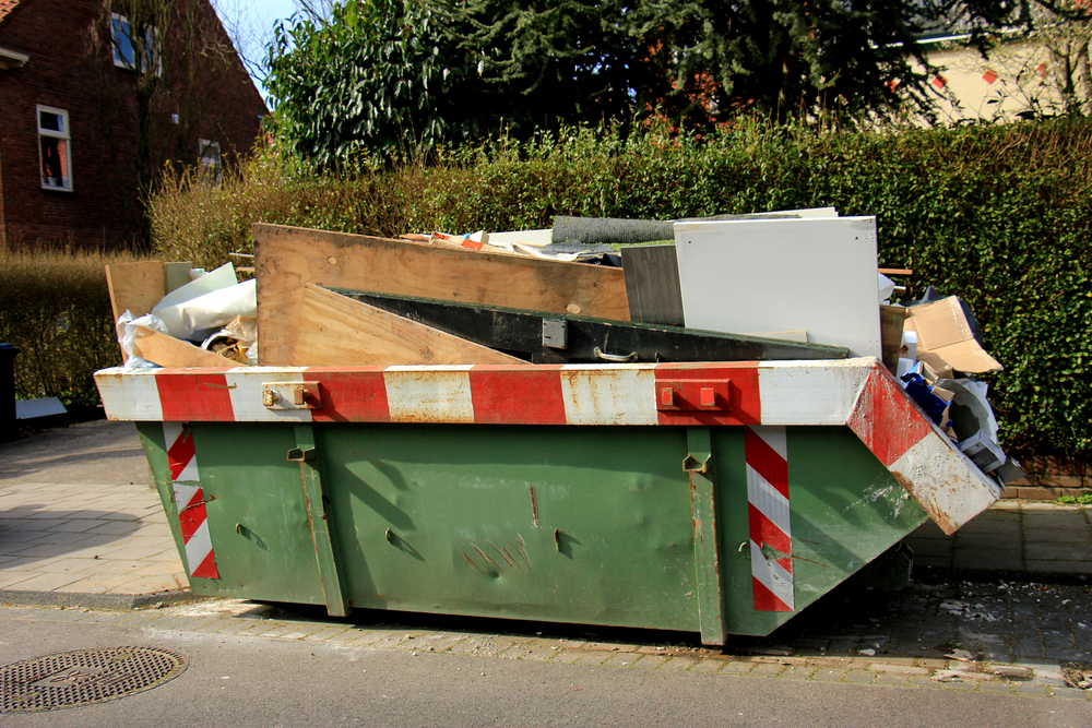 A large green skip filled with various discarded items, including wood, metal, and cardboard, sits on the side of a residential street near a hedge.