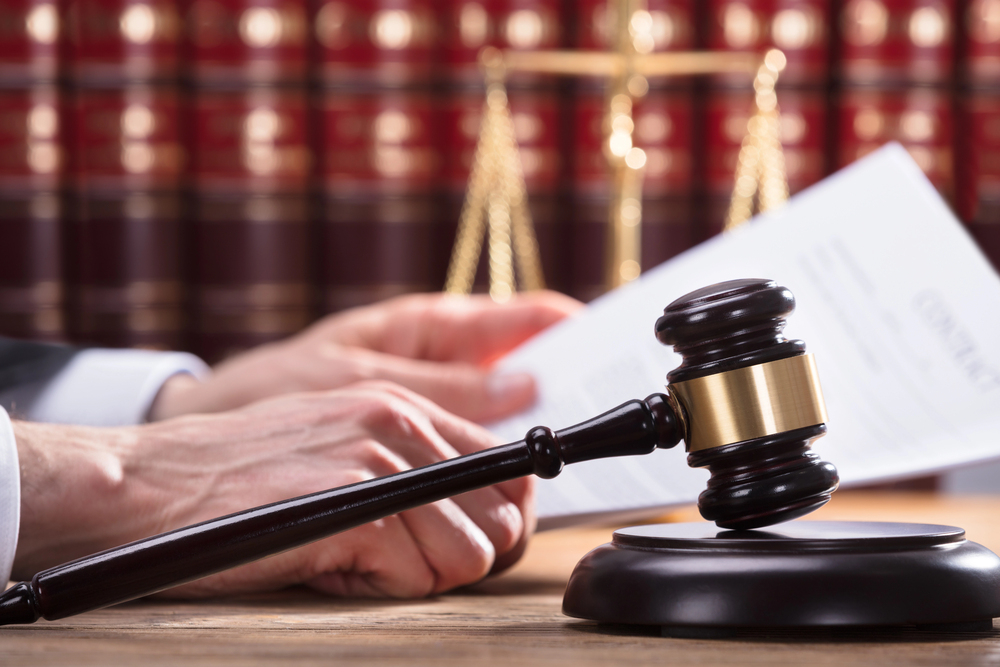 A judge's hands holding a document, with a wooden gavel and sound block in the foreground and law books and scales in the background.