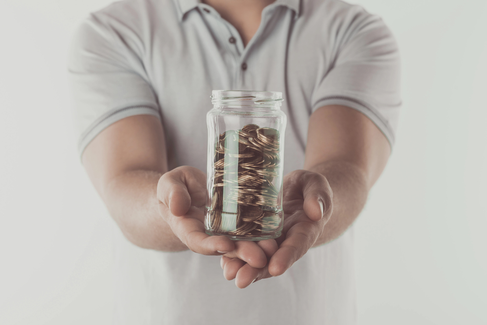 A person in a light grey polo shirt holds a glass jar filled with coins, presenting it forward with both hands against a plain white background.