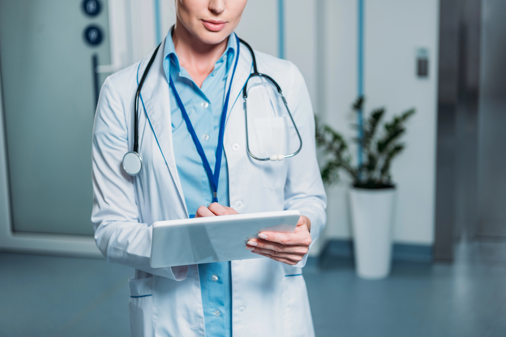 A doctor in a white coat with a stethoscope around their neck is holding and writing on a digital tablet in a medical surgery. The doctor's face is partially out of frame.