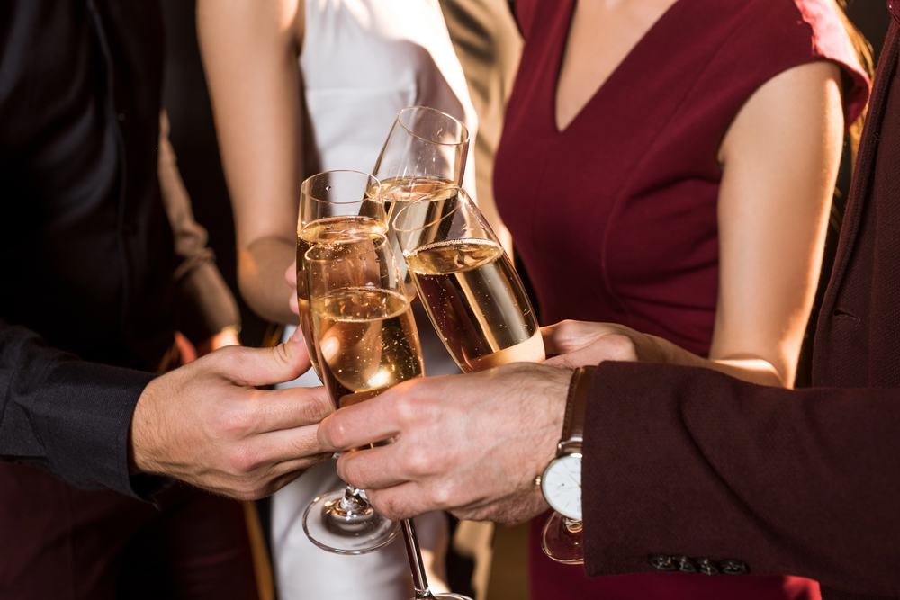 Four people clinking champagne glasses together in a celebratory toast, dressed in formal attire. The focus is on their hands and glasses, with only their torsos and arms visible.
