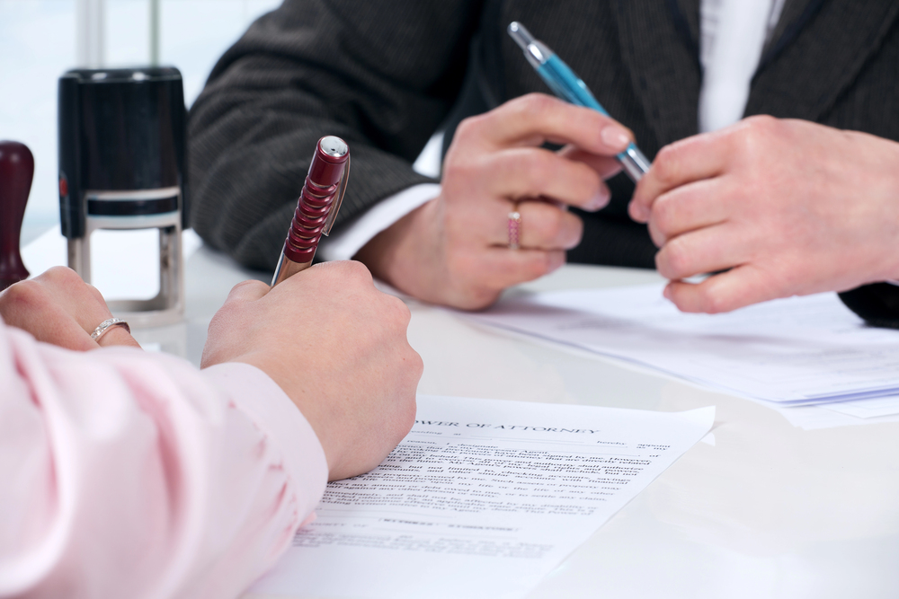 Two people sit at a desk holding pens, discussing and reviewing a power of attorney document. Stamps and paperwork are on the table. Only their hands and part of their torsos are visible.