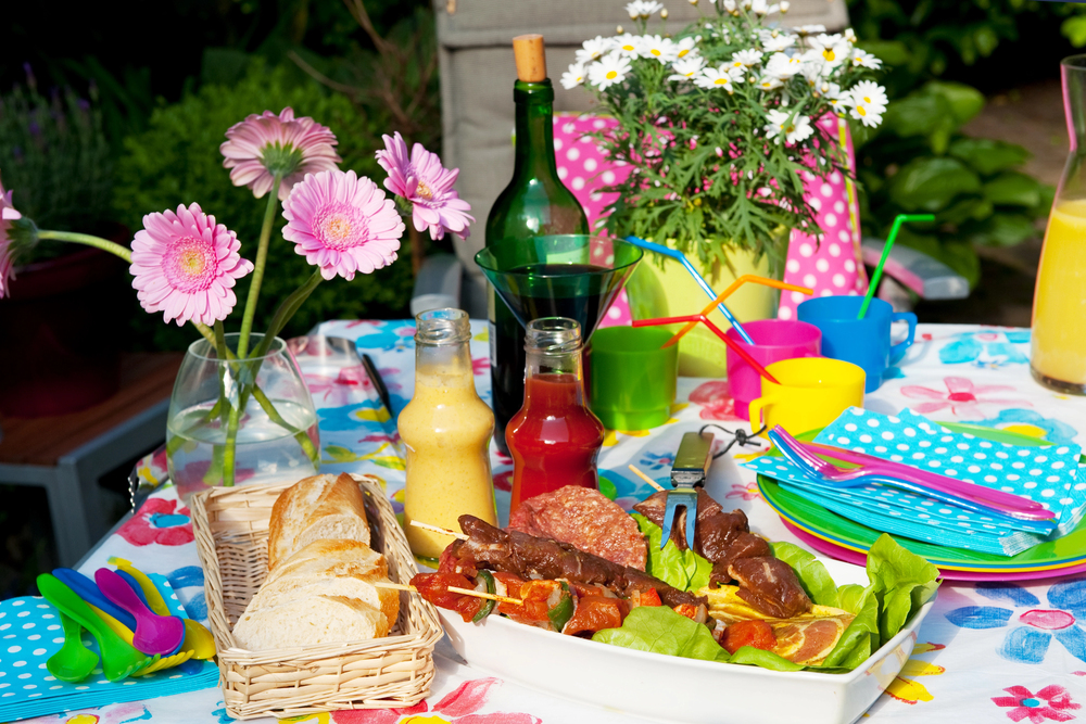 A colourful outdoor table set with flowers, bread, skewers, drinks, sauces, cups with straws, and serviettes, creating a festive picnic atmosphere in a garden setting.