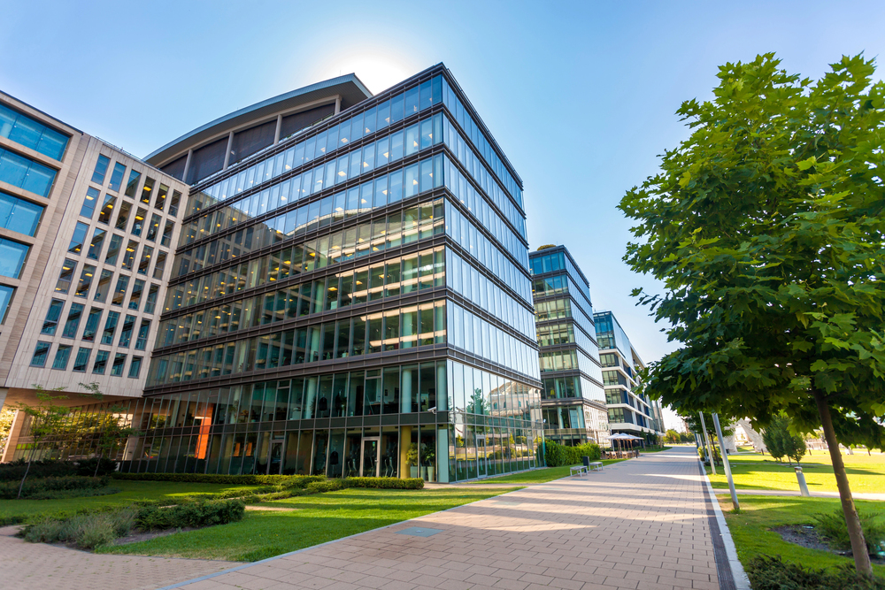 Modern glass office buildings lined along a walkway with green lawns, trees, and clear blue sky in the background.