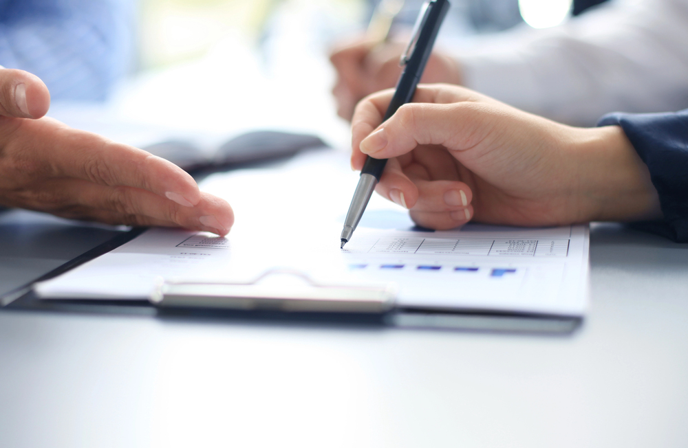 Close-up of two people discussing paperwork at a desk, one holding a pen and pointing at a document on a clipboard, with graphs and charts visible.