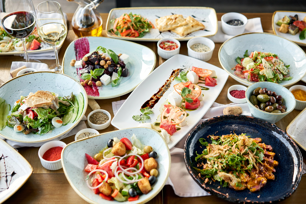 A table filled with assorted Mediterranean dishes, salads, bread, olives, and glasses of white wine, arranged on plates and bowls for a vibrant shared meal.