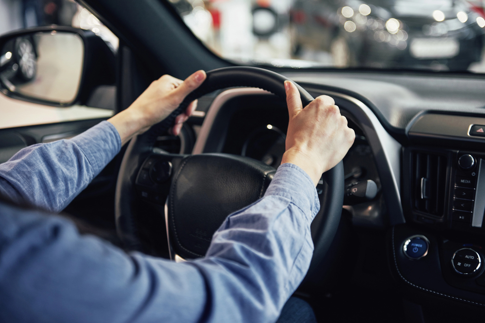 A person wearing a blue long-sleeve shirt is holding a car steering wheel with both hands, sitting in the driver’s seat. The dashboard and interior controls of the vehicle are visible.