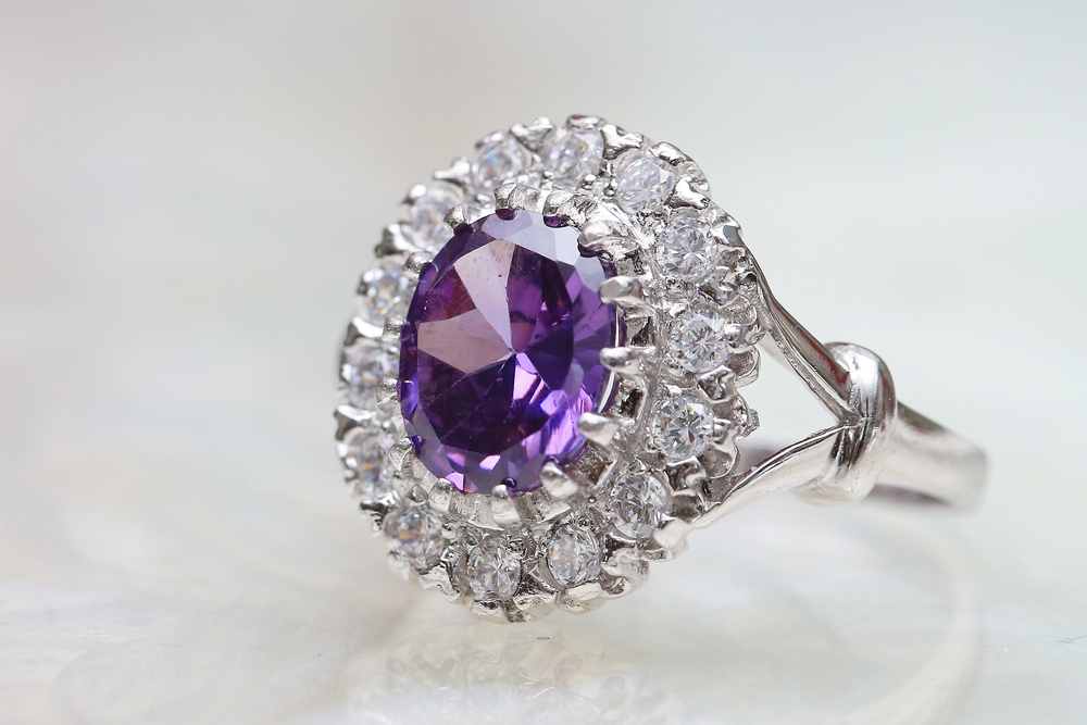 A silver ring featuring a large oval purple gemstone in the centre, surrounded by a halo of small clear diamonds, set against a blurred light background.