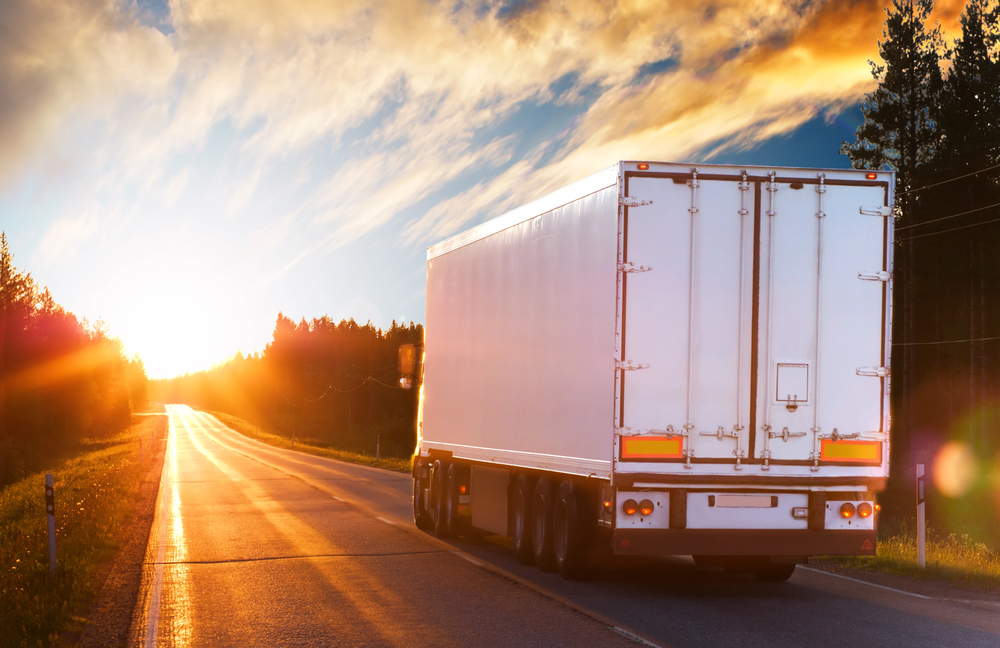 A white lorry drives down a rural road at sunset, with sunlight shining brightly and trees lining the roadway.