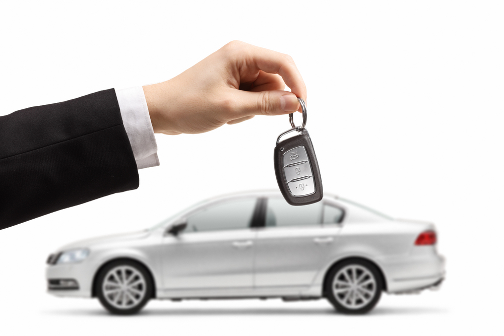 A hand in a black suit holds a car key fob in front of a silver saloon, isolated on a white background.