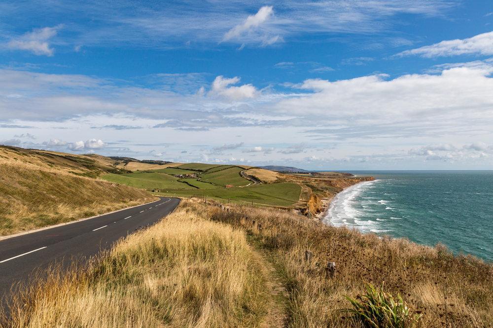 A winding coastal road runs alongside grassy cliffs and farmland, with waves crashing against the shore under a partly cloudy blue sky.