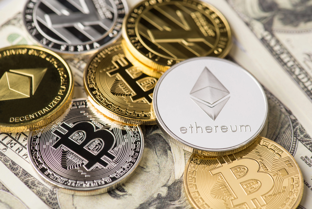 Various physical coins representing cryptocurrencies like Bitcoin and Ethereum are scattered on top of U.S. dollar notes.