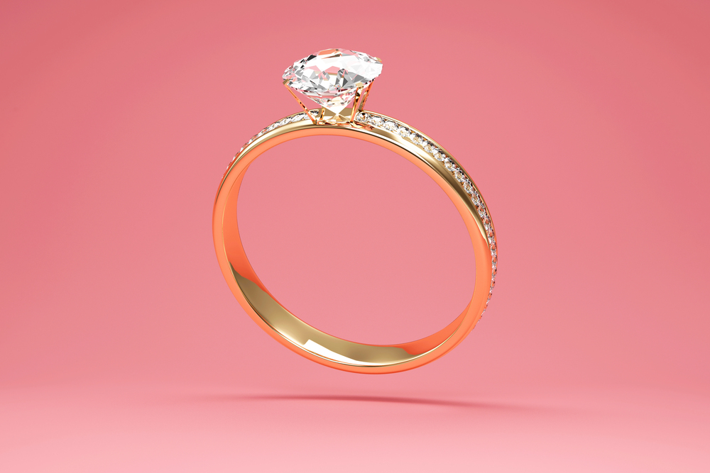 A gold engagement ring with a large, round diamond solitaire and small diamonds on the band, displayed against a soft pink background.