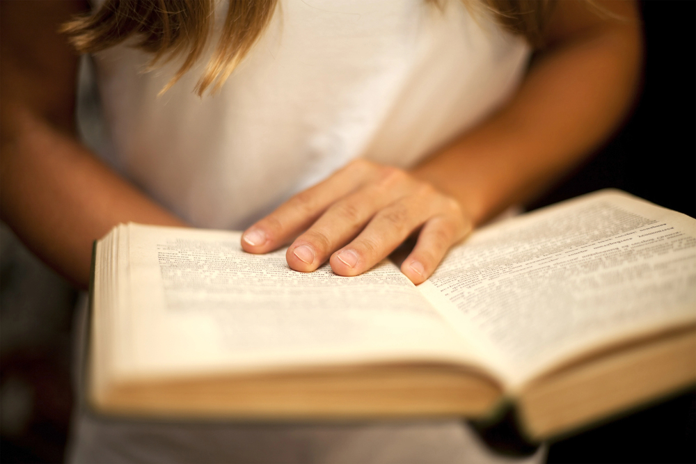 A person wearing a white sleeveless top is reading a book, with one hand gently resting on an open page.