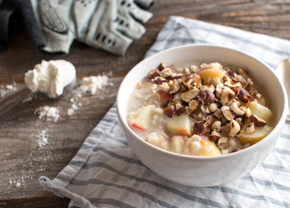 A bowl of porridge topped with chopped nuts and apple slices sits on a striped cloth, with a spoonful of flour and a pair of gloves nearby on a wooden surface.