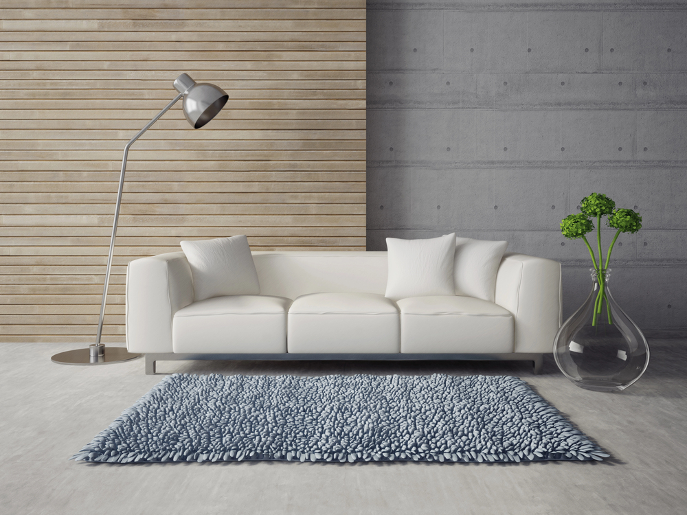 A modern living room with a white sofa, three white cushions, a silver floor lamp, a blue textured rug, and a clear vase with green flowers against wood and concrete walls.