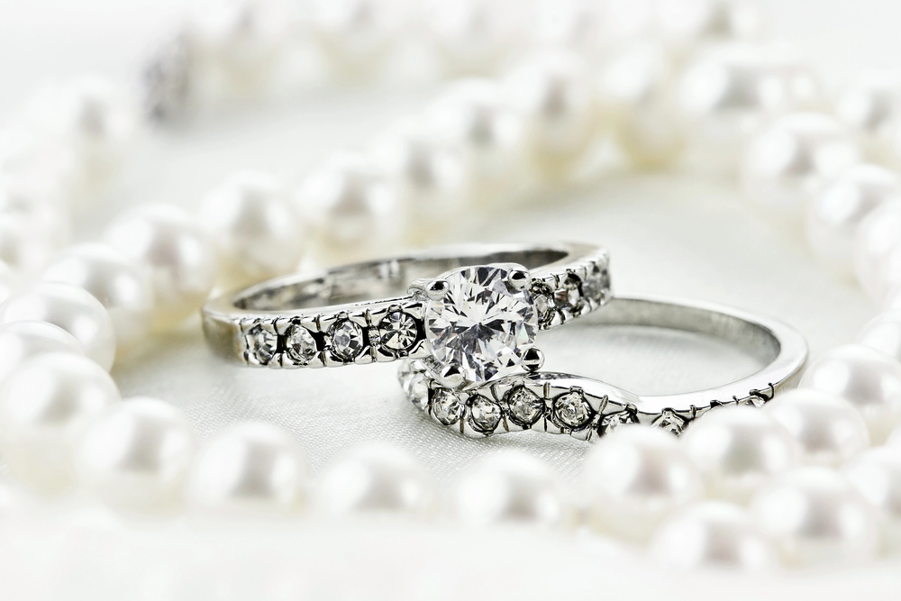 Two diamond rings, one with a large central stone, rest on a white surface surrounded by a string of white pearls.