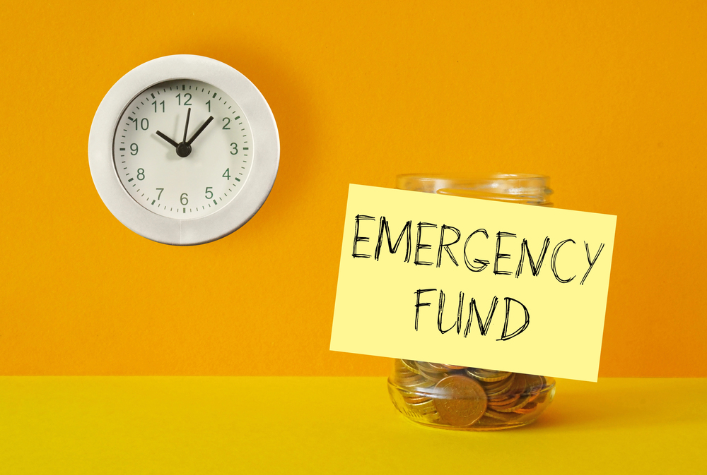 A wall clock and a glass jar filled with coins sit against a yellow background. A yellow sticky note labelled EMERGENCY FUND is attached to the jar.