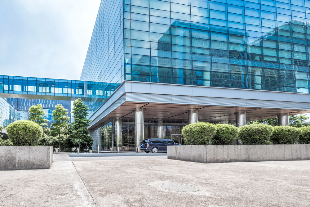 A modern glass office building with reflective windows, a covered entrance supported by columns, and a dark van parked nearby. Neatly trimmed bushes line the front and a skybridge connects to another building.
