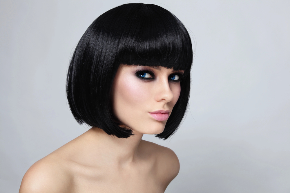 Woman with short, sleek black bob haircut, blunt fringe, and dramatic eye make-up poses against a light grey background, gazing slightly to the side with a neutral expression.